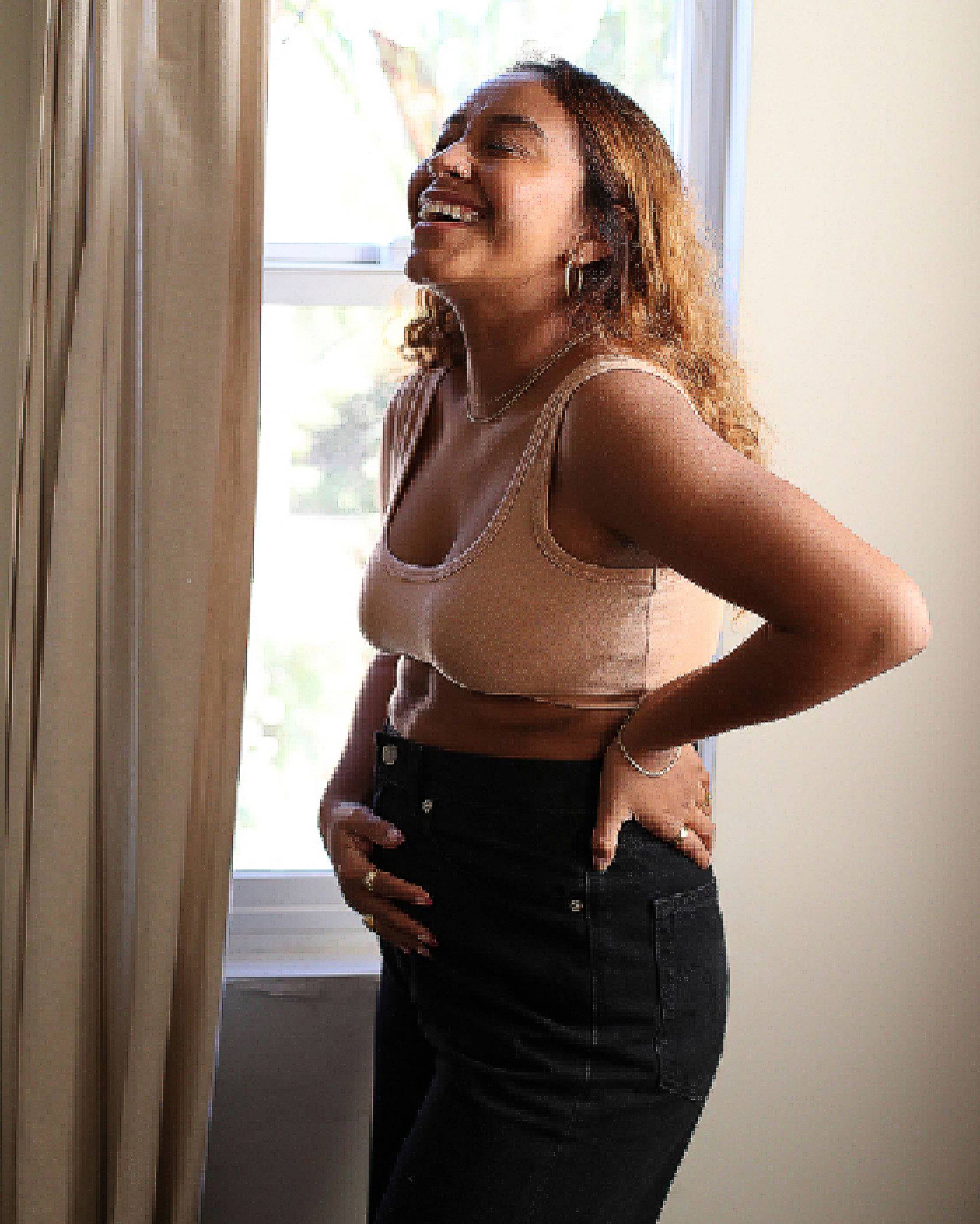 Photo of Evelynn Escobar-Thomas holding her pregnant belly and throwing her head back in laughter. She is wearing a peach sports bra and black jeans.