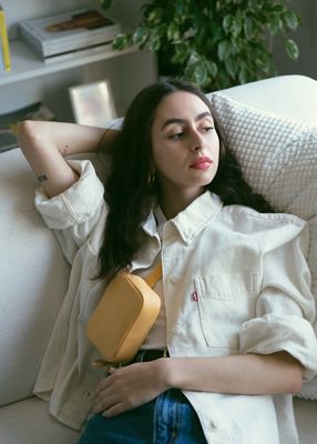 Adeline Hocine laying on a white couch while wearing a yellow belt bag and cream colored trucker jacket.