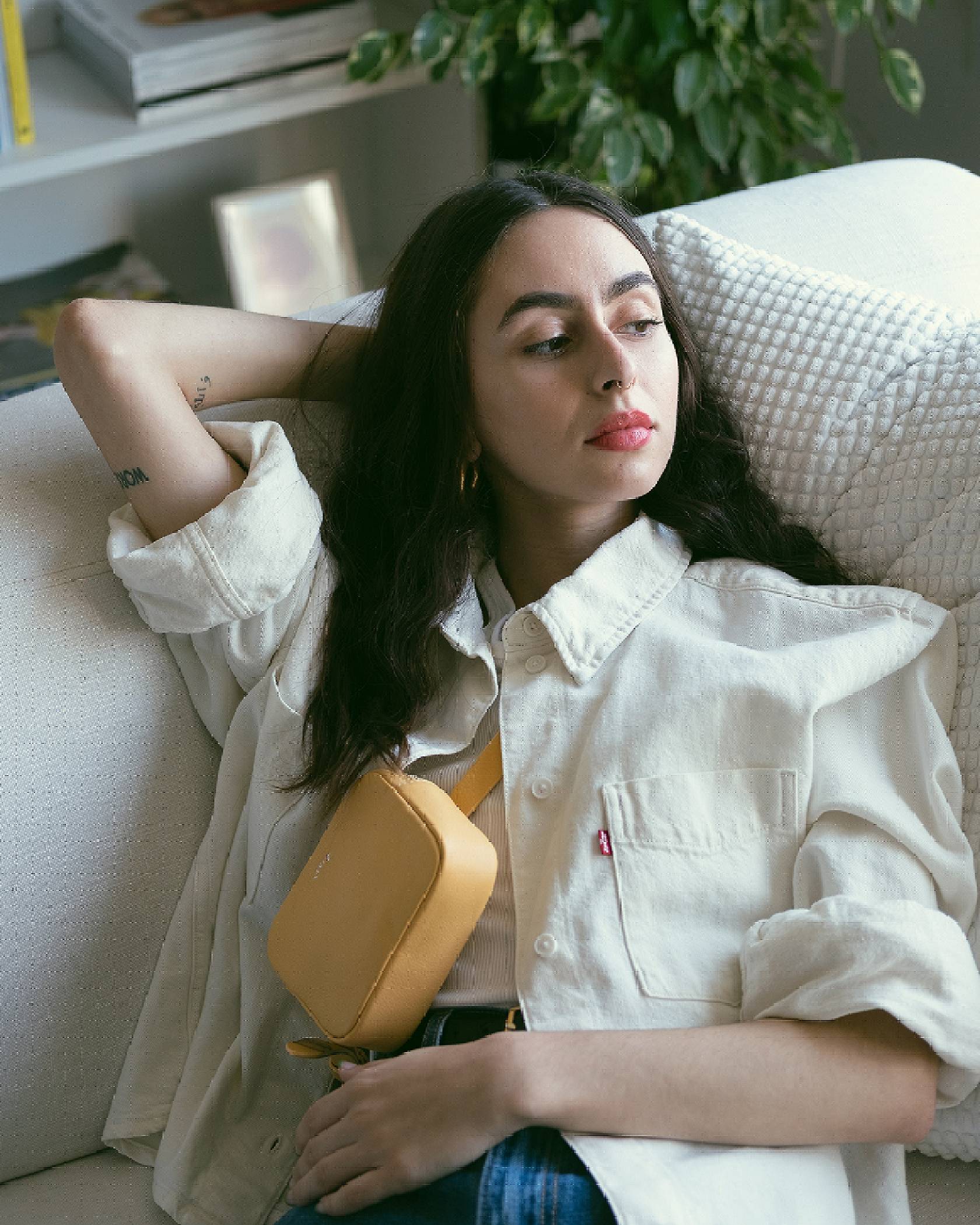 Adeline Hocine laying on a white couch while wearing a yellow belt bag and cream colored trucker jacket.