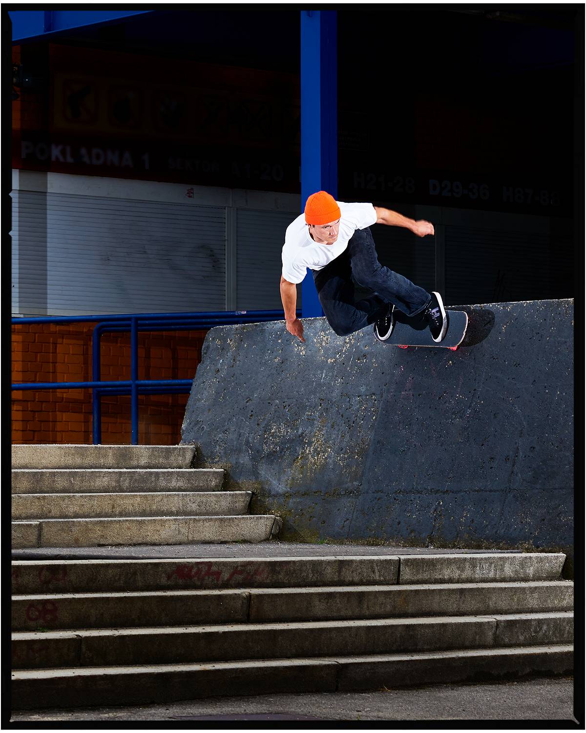 Person wearing an orange beanie, white tee, and Levi's jeans while skateboarding on a wall by a staircase.