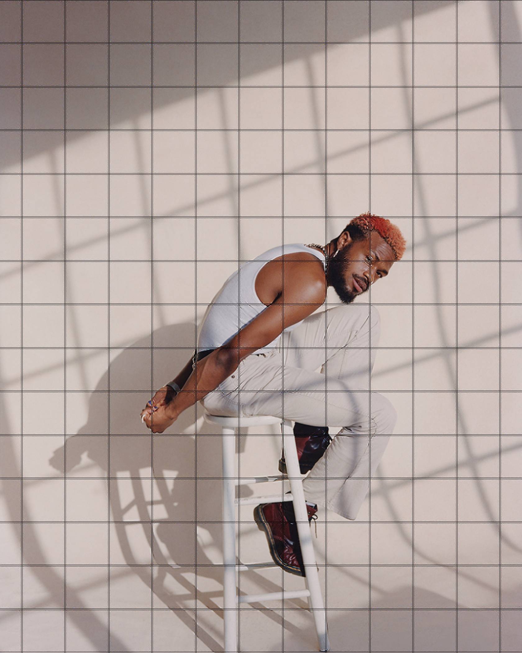 Photo of Duckwrth sitting on a white stool wearing a white tank top and white jeans with black boots.