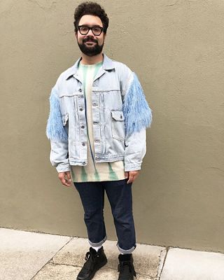 Photo of Max Kessler standing in front of a tan wall. He is wearing thick black eyeglasses, a white and turquoise sweater, a Trucker Jacket with blue fringe on the sleeves, cuffed Levi's jeans, and black sneakers.