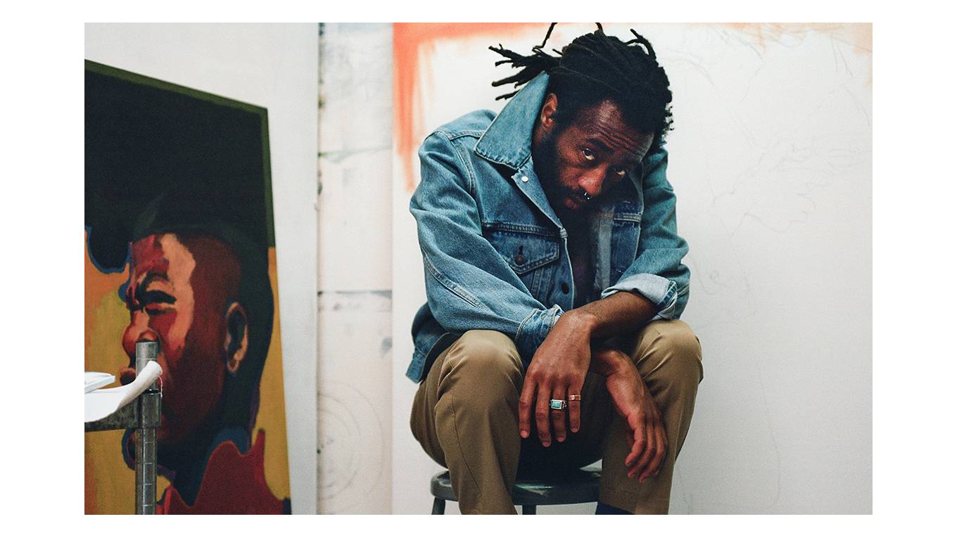 Portrait of Joonbug sitting in his studio next to a painting.