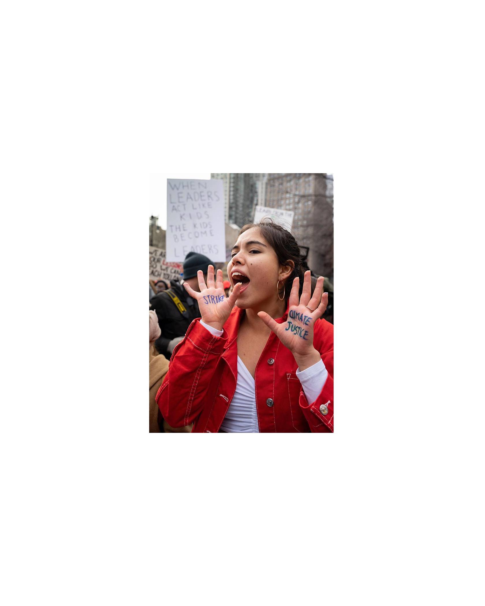 Photo of Xiye Bastida with her palms raised to face the camera. One palm reads "CLIMATE JUSTICE" while the other palm reads "STRIKE" in black marker. She is wearing a white top and a red jacket.
