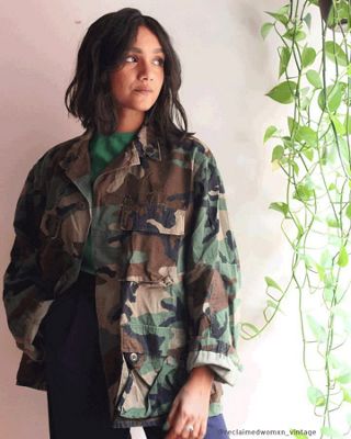 GIF of Tahia Islam standing next to a hanging plant in a green shirt, camo jacket, and navy pants.