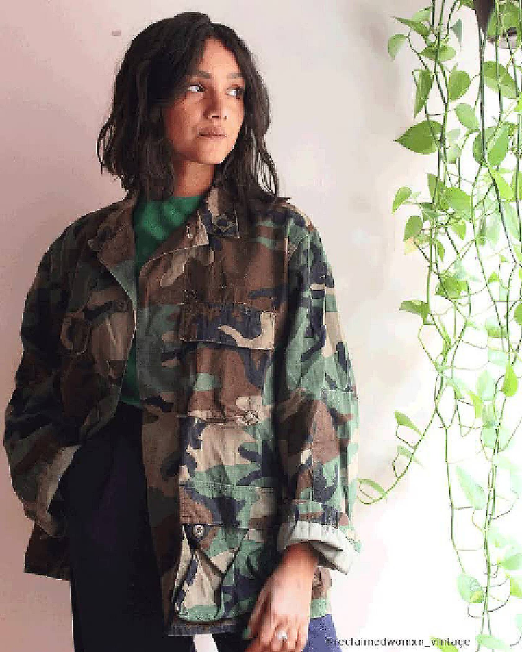 GIF of Tahia Islam standing next to a hanging plant in a green shirt, camo jacket, and navy pants.