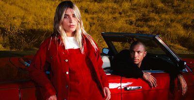Woman in red overalls leaning on car with man sitting in car in the background.