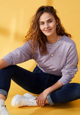 Image of model Elena Carrière sitting on the floor that has a yellow background, wearing a purple sweatshirt, dark wash denim jeans, and white shoes while her left arm holds her left ankle.