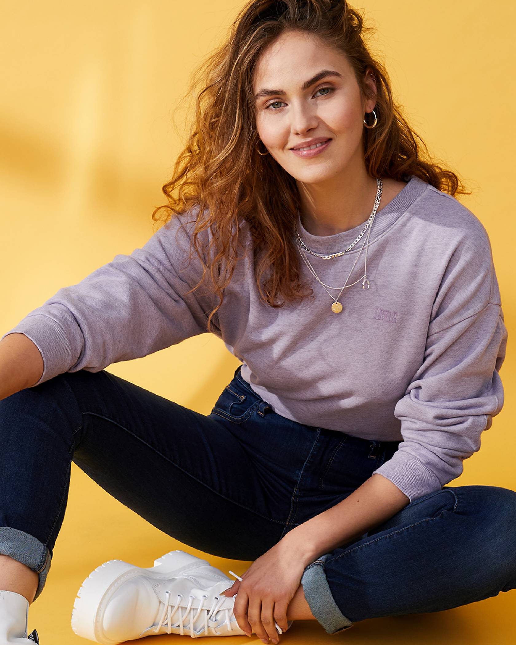 Image of model Elena Carrière sitting on the floor that has a yellow background, wearing a purple sweatshirt, dark wash denim jeans, and white shoes while her left arm holds her left ankle.