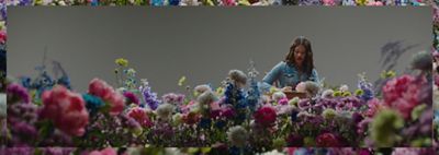 Image of Leyna Bloom sitting at a wooden desk, wearing a denim western shirt, writing on a stack of papers while surrounded by colorful flowers.