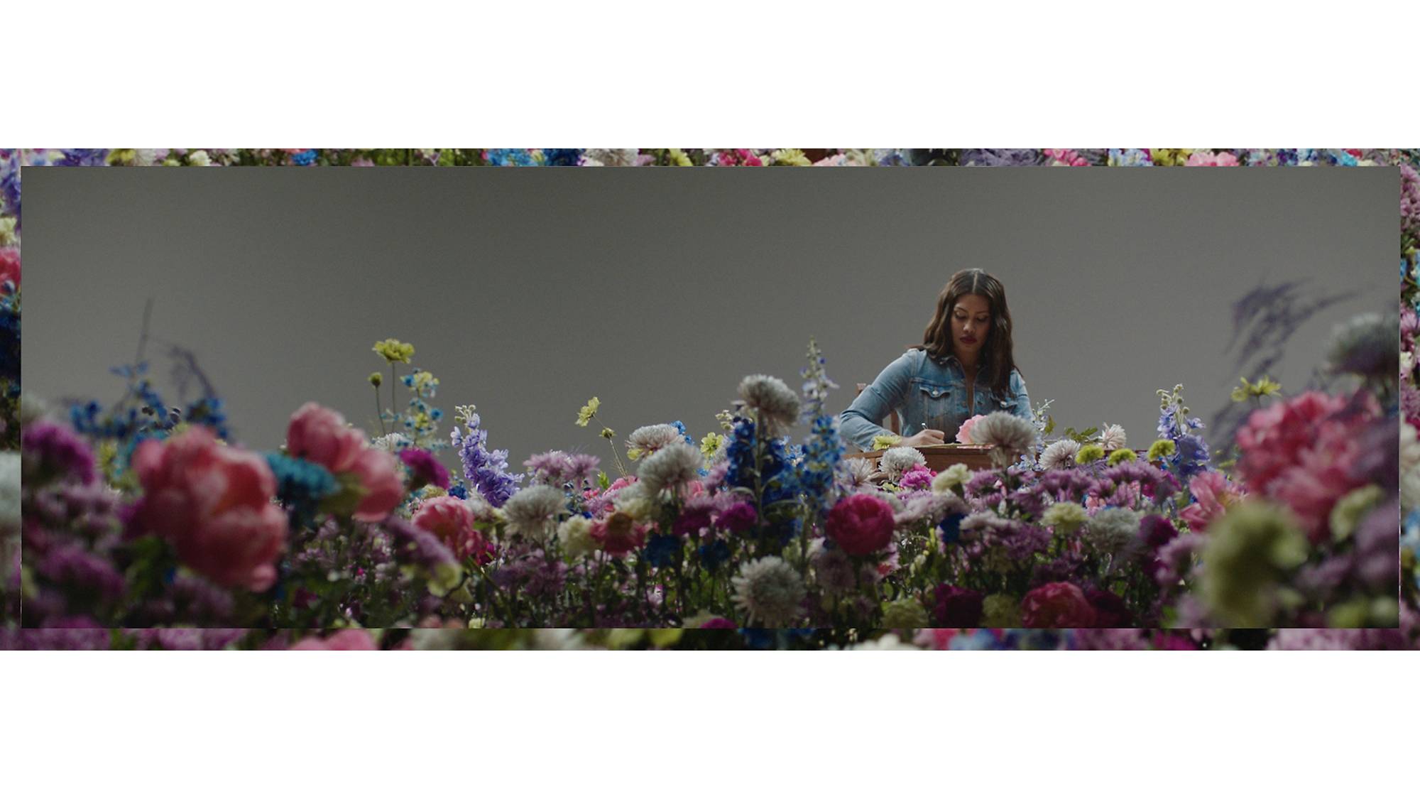 Image of Leyna Bloom sitting at a wooden desk, wearing a denim western shirt, writing on a stack of papers while surrounded by colorful flowers.